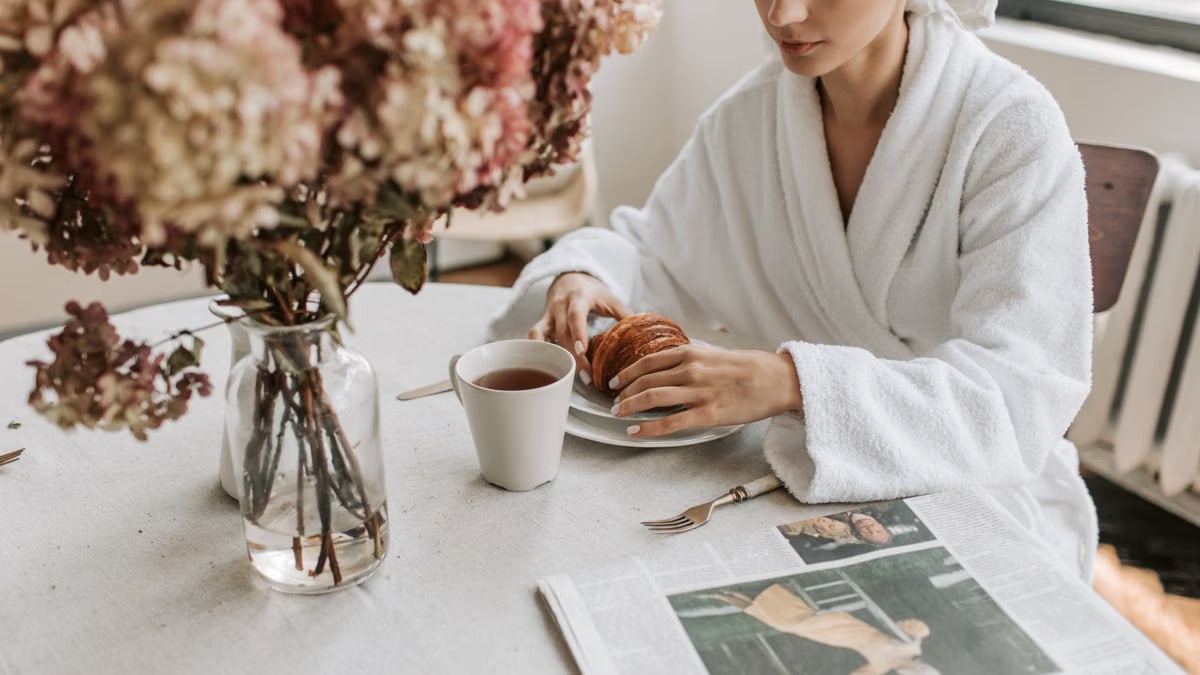 A woman in a bathrobe having breakfast indoors with a croissant and tea.